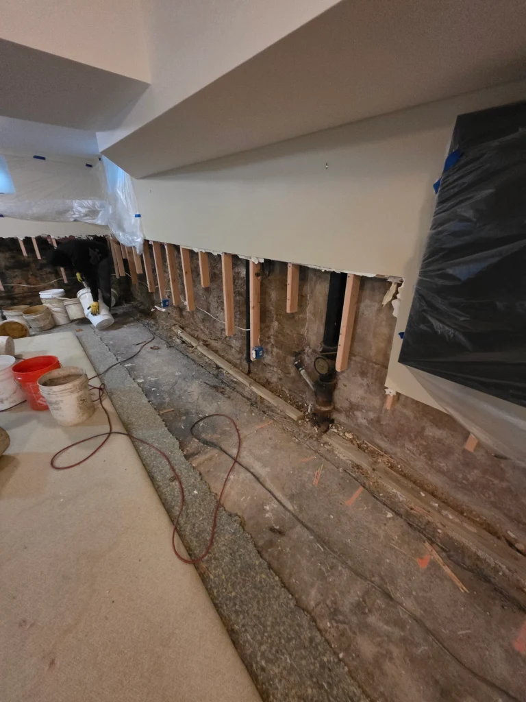 Wall construction scene with exposed wooden studs, pipes, and partially applied drywall. Buckets and tools scattered, conveying work in progress.