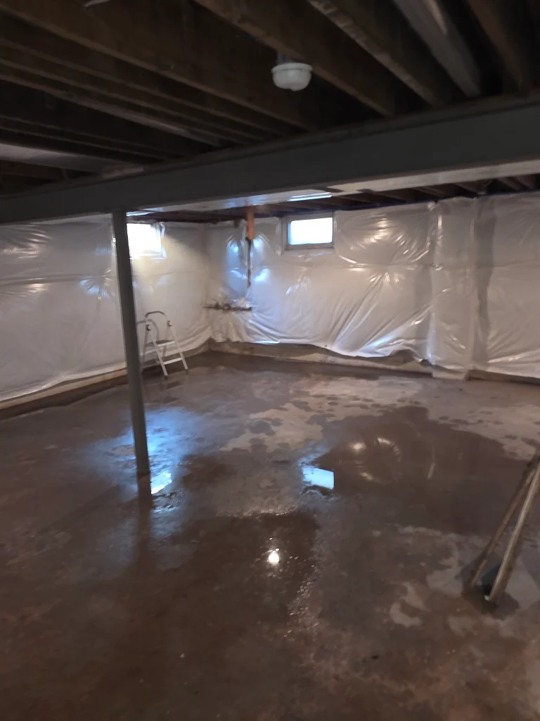 Basement with concrete floor showing water puddles, white plastic-covered walls, exposed ceiling beams, a smoke detector, and a metal support beam.
