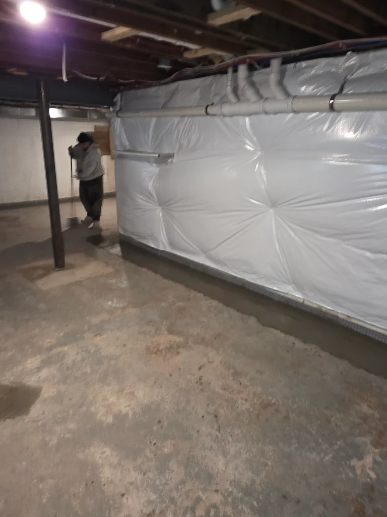 A person mops the floor in a dimly lit basement with exposed beams. Walls are covered with white insulation, and pipes run along the ceiling.