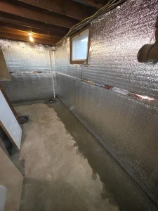 A dimly lit basement with exposed wooden beams and reflective insulation on walls. A small window lets in light. The concrete floor has wet patches.